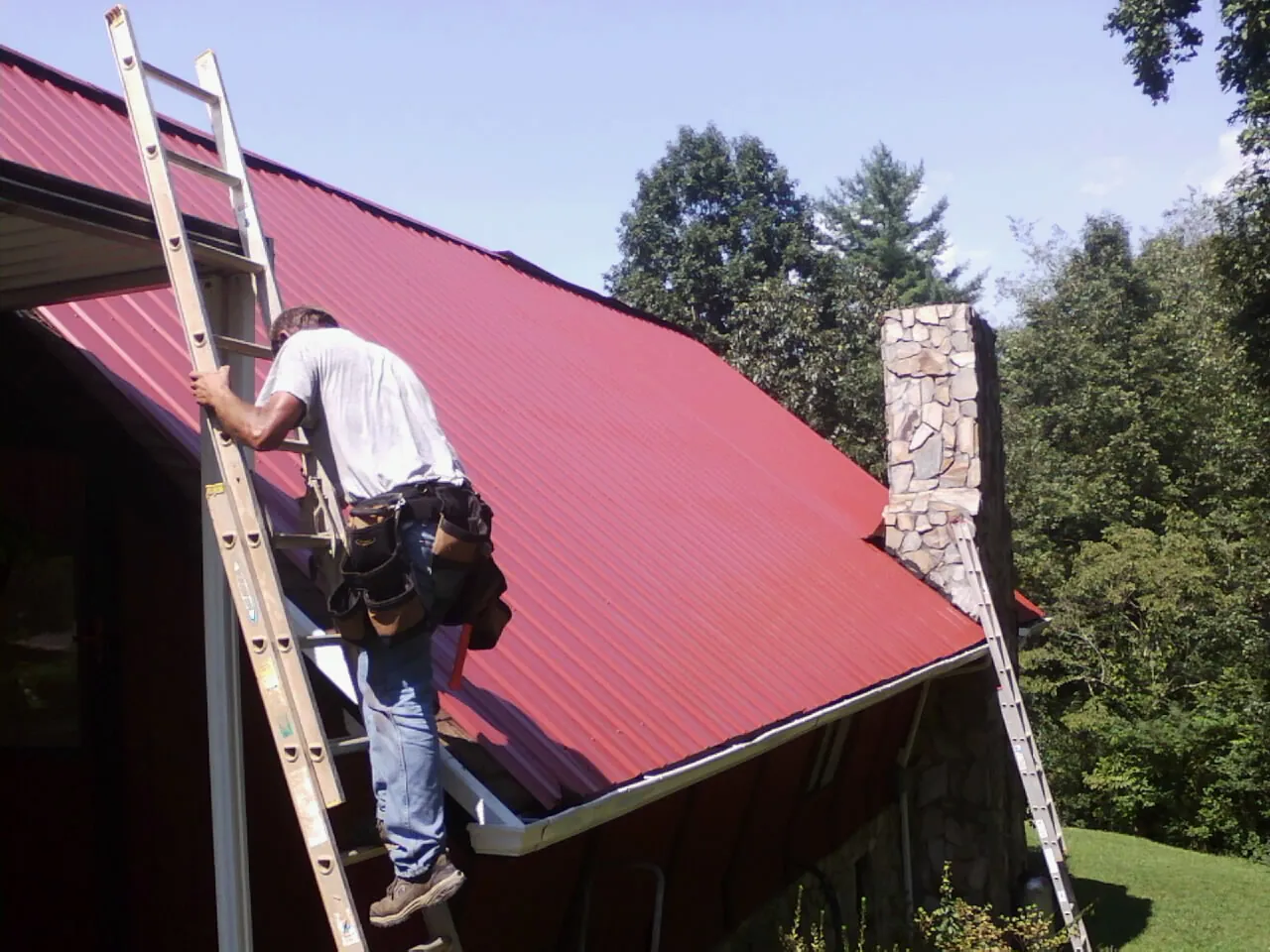 Roofer on a ladder beside a red metal roof and stone chimney — Soffit & Fascia Repair by Roofing Brothers in Ypsilanti
